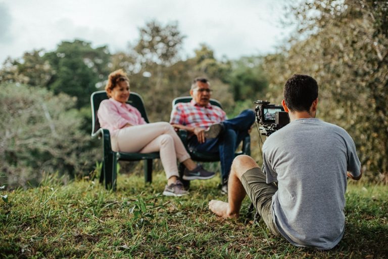 Pesquisa nacional vai mapear como turistas escolhem destinos, transportes e hospedagens Casal sendo entrevistado ao ar livre, em um jardim arborizado.