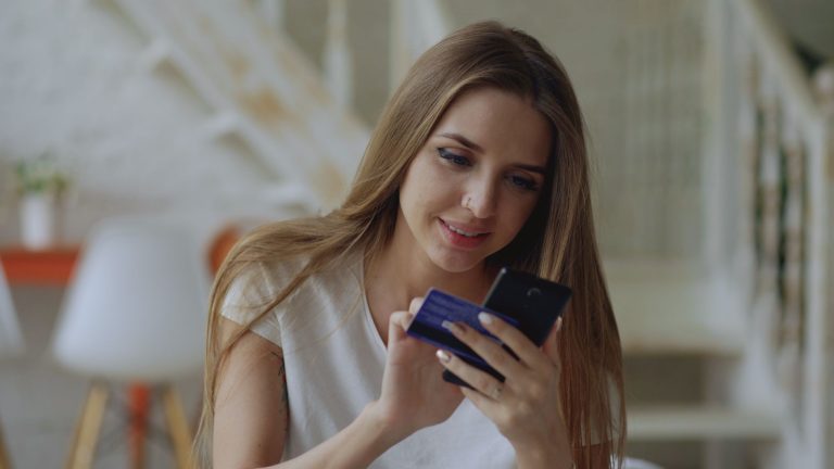 Mulher usa um smartphone para fazer uma compra online enquanto segura um cartão de crédito em casa.